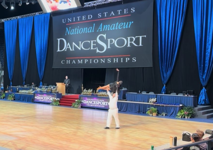 Competitive ballroom dancers performing at a national DanceSport event under stage lighting
